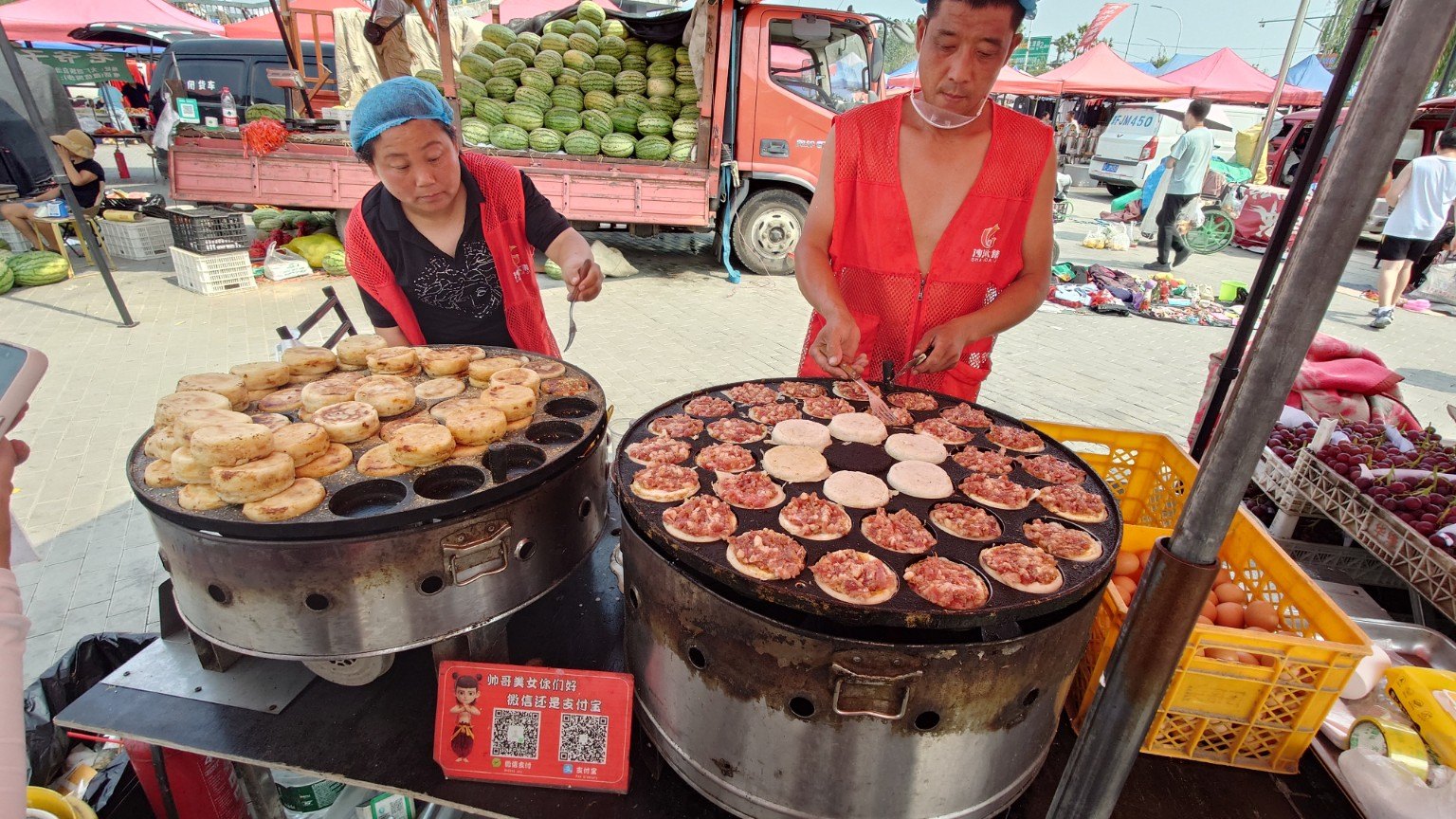 Two vendors cook meat-and-egg burgers on large round iron griddles beside a truckload of watermelons