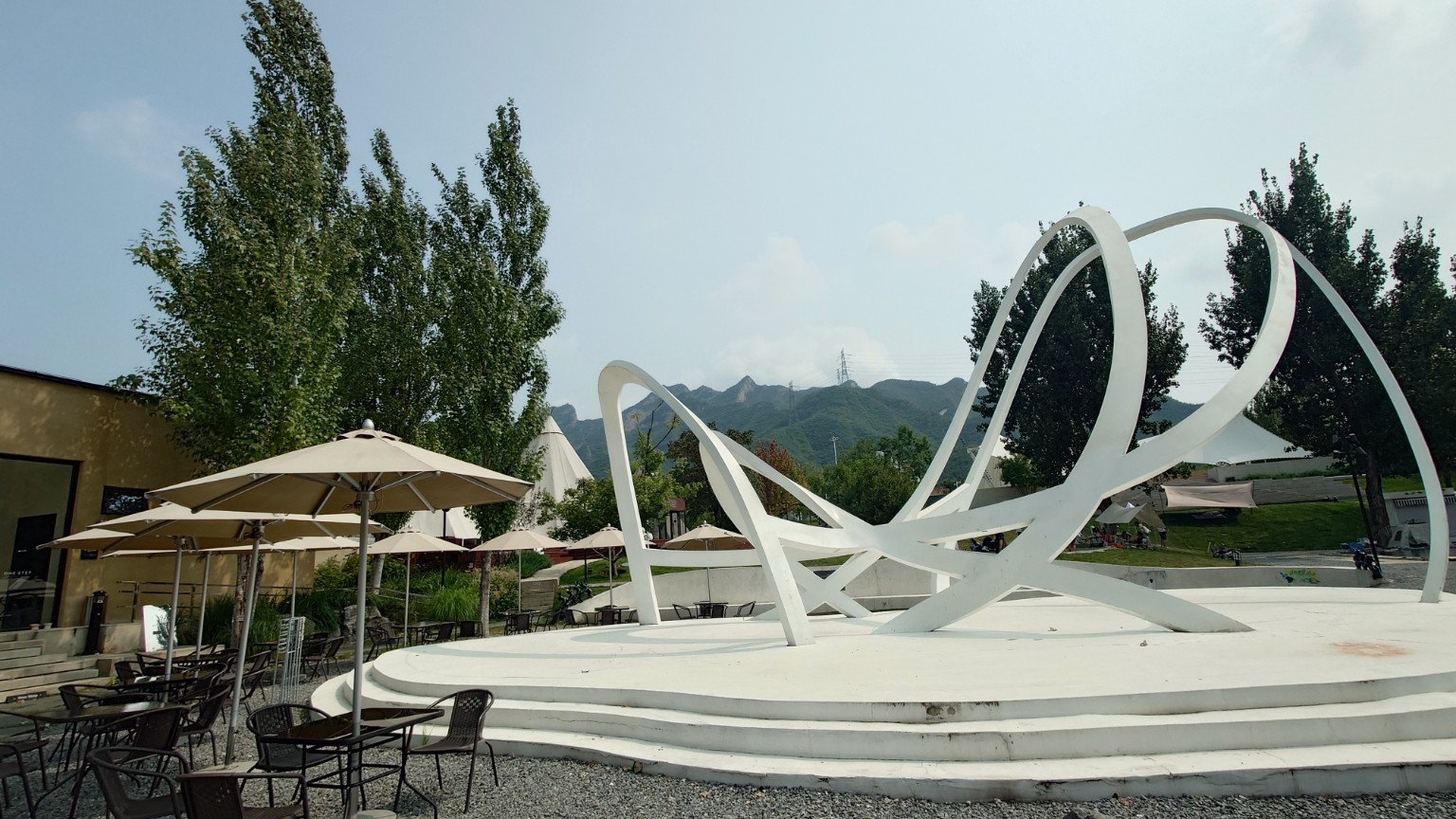 A white abstract sculpture surrounded by café seating with Tanzhesi’s mountains behind
