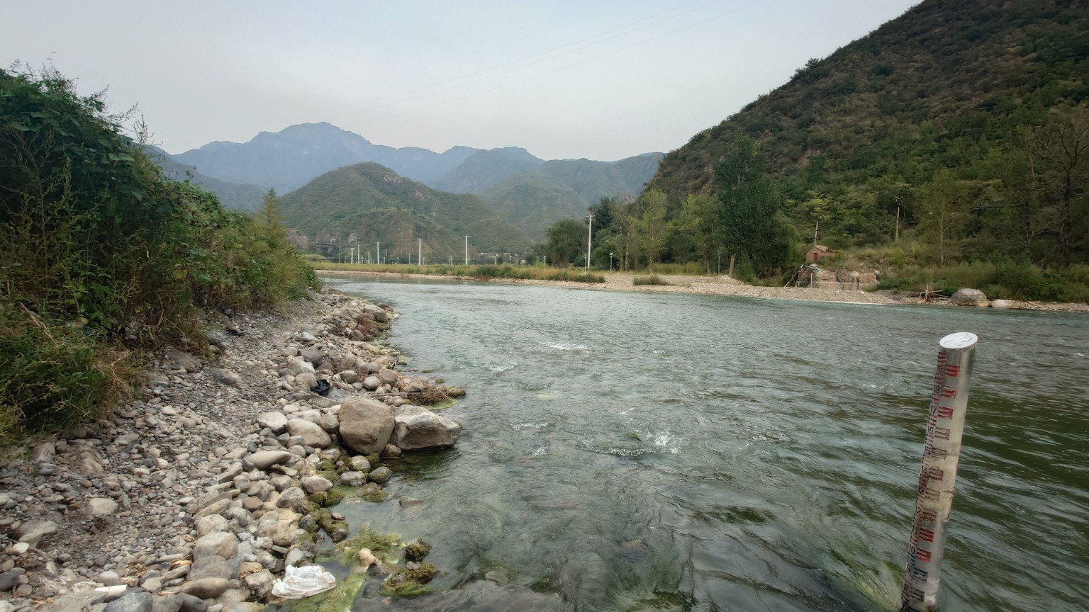 Flowing Yongding River water beside a stony bank and water-level gauge, with mountains rising on both sides