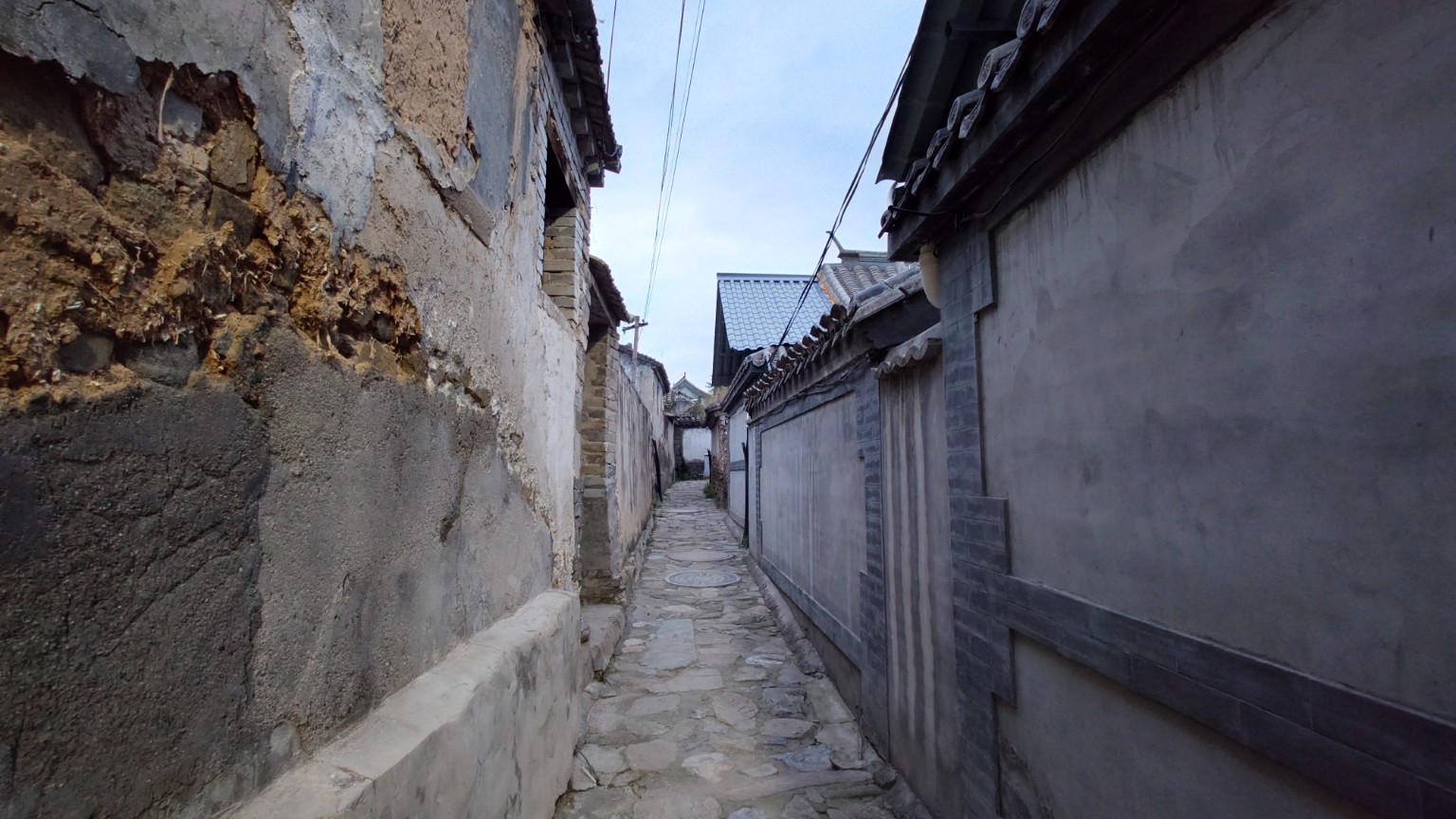 An old earthen wall facing a repaired grey-brick courtyard across a narrow stone lane