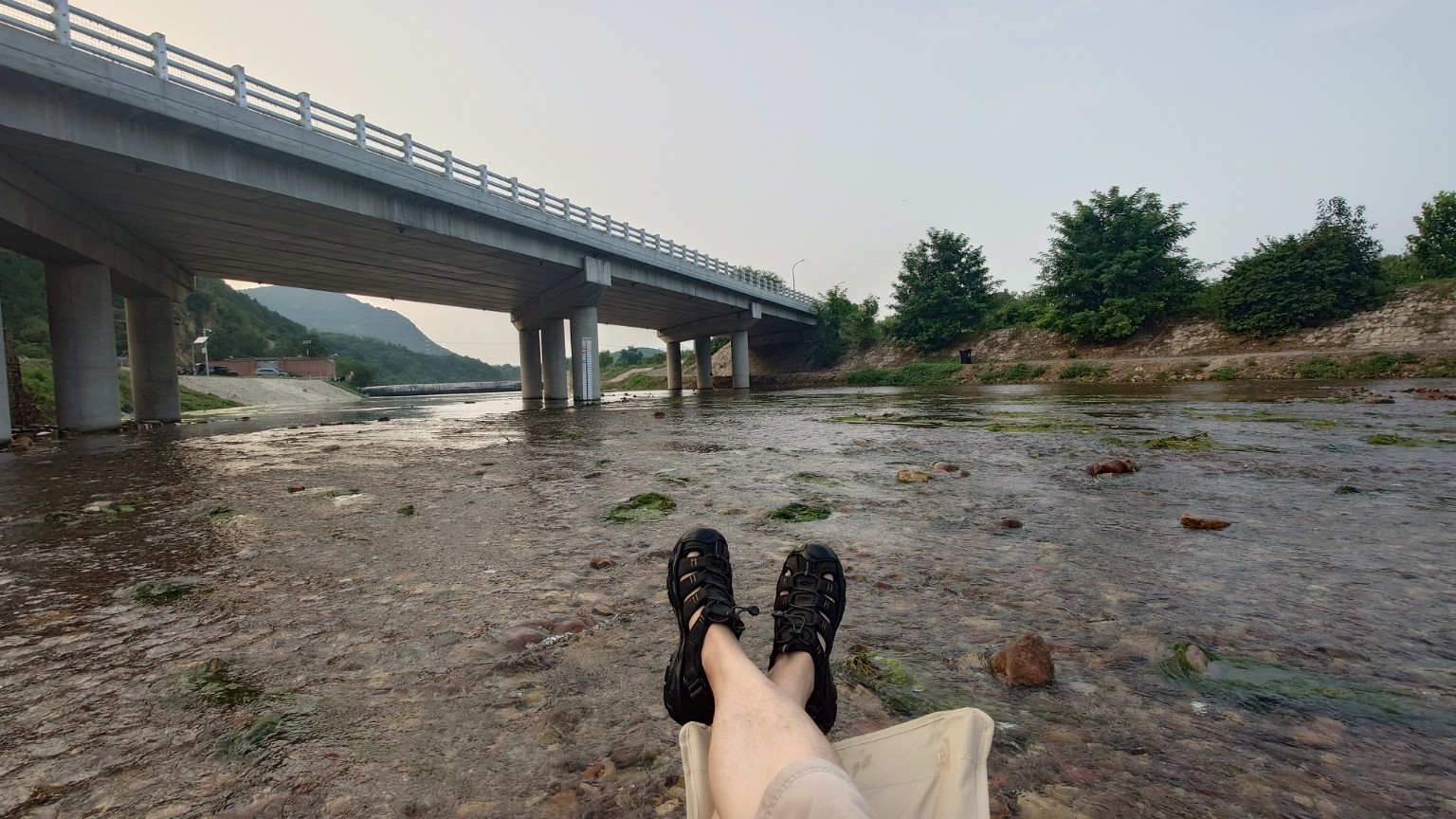A person resting in a folding chair in a shallow river beneath a bridge in Pinggu