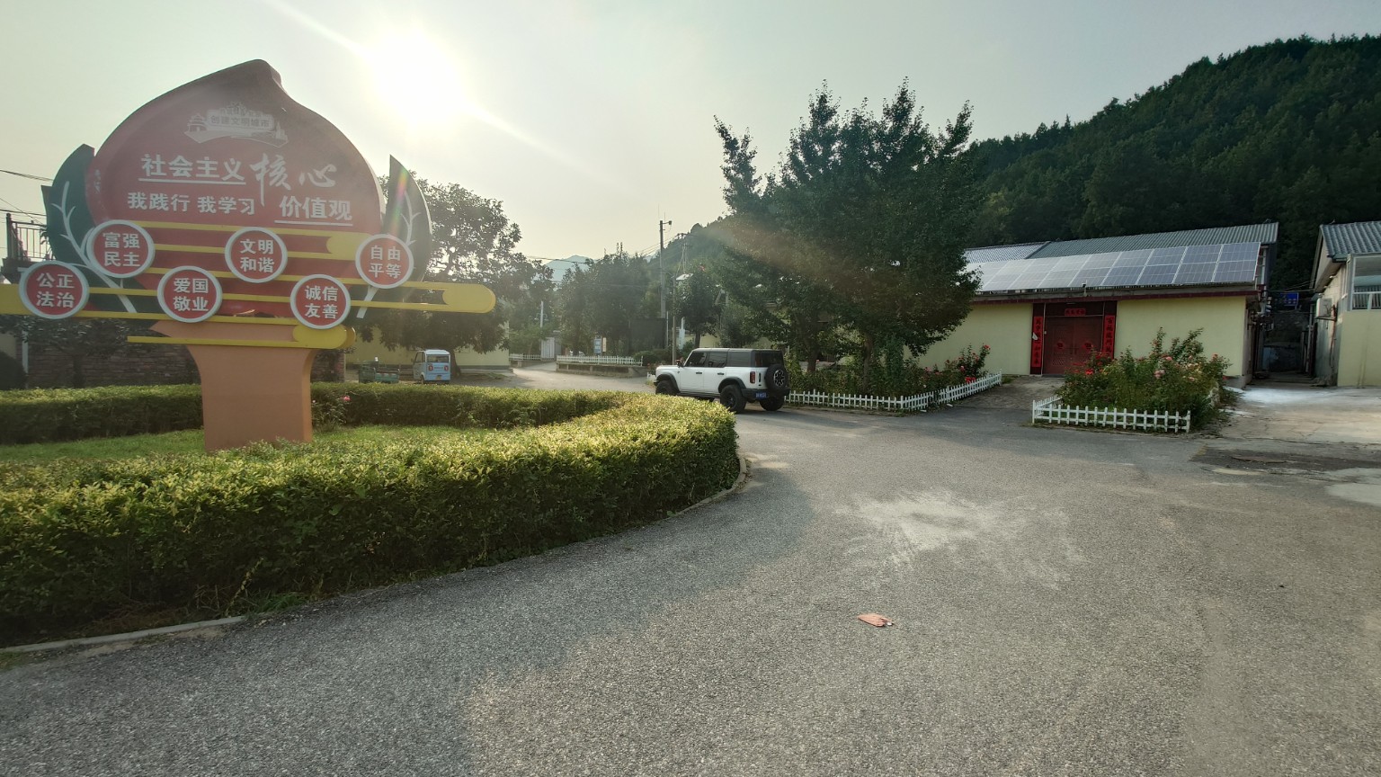 A clean village street with hedges, courtyard homes and wooded hills in late-afternoon light